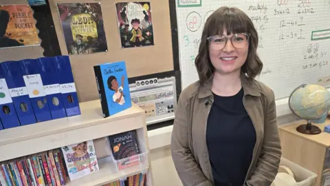Lucie Bebbington-Lewis has brown hair in a short bob. She has on glasses and has pale skin. She is standing in a classroom. 