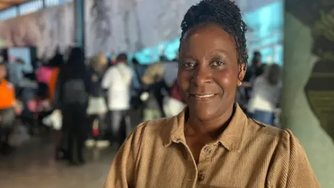 Jon Wright/BBC Mollin Delve stands smiling at the camera. There are people behind inside a conference room.