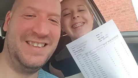 Les Sutton  Anna smiles as she holds up her A-level results, printed on a white sheet of paper. She is sat next to her dad, Les Sutton, in a car