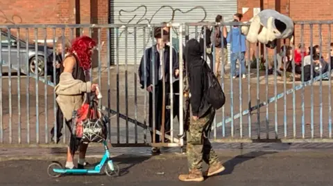 People around a warehouse, some dressed in fancy dress costumes, one person is riding a scooter. Their faces have been pixelated.