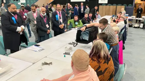 Shariqua Ahmed/ BBC A man emptying a black square ballot box onto a table lined with counters, while people on the other side of the table are watching the papers fall on the table.