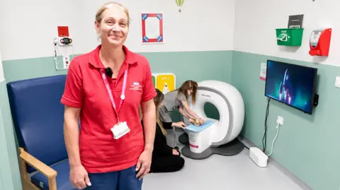 Sonia Dugmore is a senior health play specialist at the John Radcliffe. She is wearing a red polo shirt. Behind her, Ashley and Macie are using the kitten scanner.