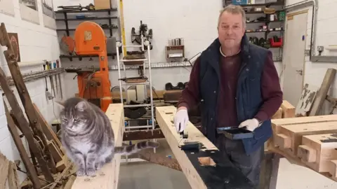 Supplied Howard Stafford is painting a wooden plank in black while restoring the Hetton Chaldron Wagon in a workshop. He is wearing a red jumper, blue body warmer and and black trousers stained with paint. There are working tools and storage shelves behind him. A fluffy grey cat is sitting on a wooden plank opposite the one which is being painted.