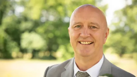 Shanice Miller A bald clean-shaven man is smiling at the camera. He is wearing a grey suit, white shirt and grey tie and the background is blurred
