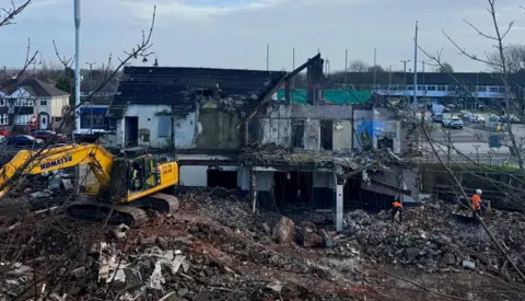 A yellow and black digger is situated in front of the half demolished former pub in Aintree