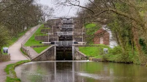 David Dixon There are five locks in a row that appear to rise in a staircase formation. In the foreground is a basin of water and there are trees and a footpath at the side of the locks. 