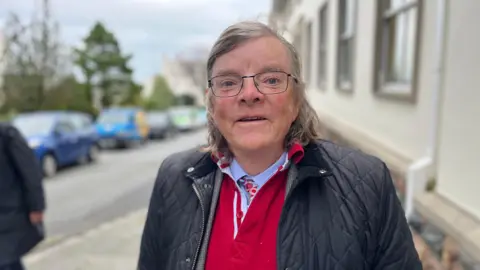 An image of Deputy Charles Parkinson. He is wearing a red shirt with a black coat on top of it. He has glasses and long grey hair.