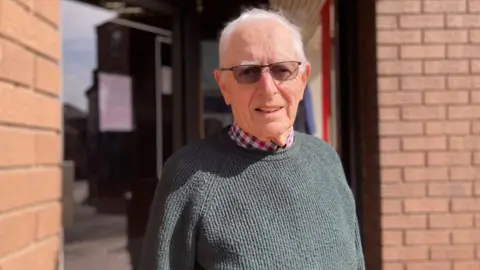 Alex Pope/BBC Ian Wood standing outside the shop with short grey hair and glasses on. He is wearing a checked shirt and green jumper. A door into the building is behind him, with brick walls either side.
