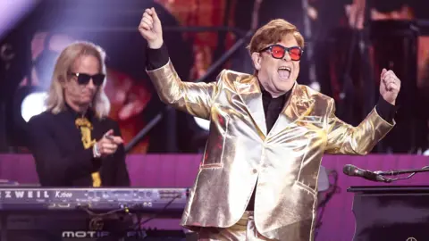 EPA Elton John, wearing a shiny gold suit and red glasses, performs at Glastonbury Festival in June 2023.