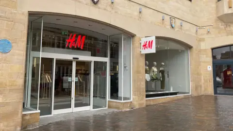 Outside of the new H&M. The building is made of light brown bricks, and has one large window and a set of white automatic doors. A H&M sign, which is red, sits above the entrance.