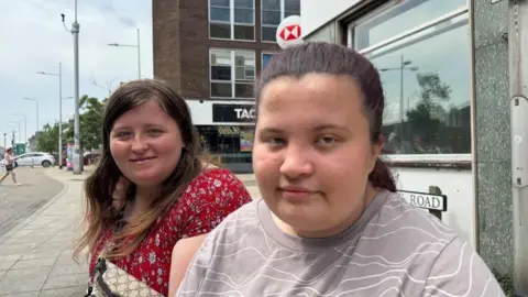 Andrew Turner/BBC Sophie is on the left of the image, with long brunette hair, and wearing a red patterned top with a bag over her shoulder. Emily is on the right, with her dark red-tinted hair tied back. She is wearing a beige top featuring a white random lines pattern. They are pictured outside the HSBC bank, where its logo can be seen above Emily's head.