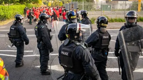 Seven police officers are in riot gear. They have helmets on and their faces are covered in a plastic shield. Football fans wearing red tops are in the background. 