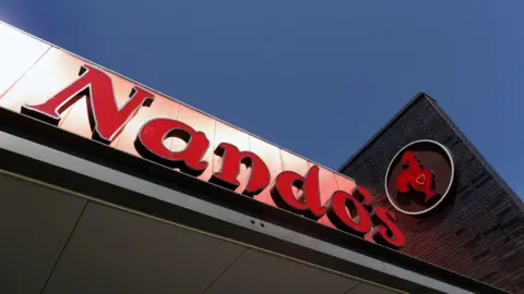 Getty Images A large Nando's sign in red lettering on a black background is seen on a sunny day with blue skies. The company's chicken logo is also visible