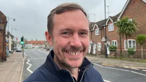 Andrew Turner/BBC Adam Blake, with mousy brown hair and goatee beard, is smiling by the roadside through Acle. He is wearing a blue jacket.