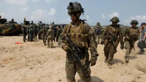 Marines dressed in camouflage clothing, holding guns, walk down a sandy beach.