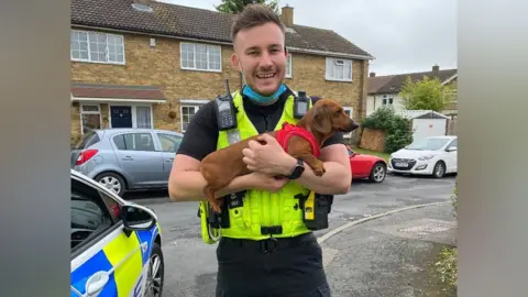 Linkedin A police officer, young, is wearing a uniform and holding a puppy next to a police car on a residential street