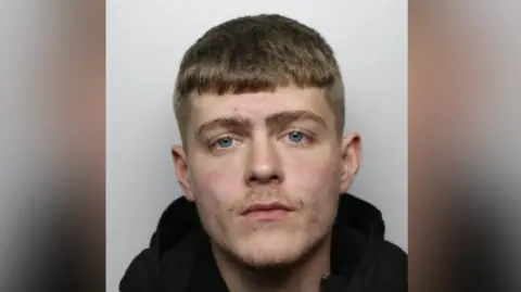 Josh Mansfield custody picture. Mansfield is staring at the camera. He has short, brown hair and is wearing a black hooded top.