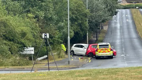 A red car is squashed in the middle of a police car and a white car. All cars are on the side of the road and pointing out into the road. A man in a hi-vis yellow jacket is sitting on the side of the road by the cars. Overgrown hedges to the right disguise a street sign that reads 'Billingham'