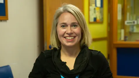 Dr Helen Munroe is smiling and has a blonde bob-cut. She is wearing a black long-sleeve top. Sat in front of a clinical background and you can see a hint of a light blue NHS lanyard around her neck