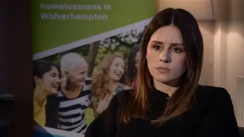 A woman with long brown hair and a black jumper looking at the person interviewing her, who is in silhouette