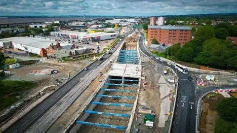 Aerial view of the site. The road is completely dug up in the middle of the carriageway with blue steel supports attached to the sides of the road as they dig down to a lower level.