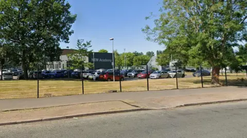 Dozens of cars parked in front of a large building situated behind gates on a sunny day.