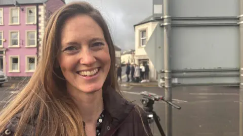 A woman smiling at the camera. She is wearing a brown coat. Behind her the back of a road sign. Across the road, also behind her, some buildings and some people walking on the footpath.