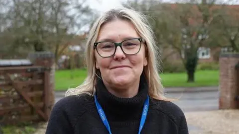 Shaun Whitmore/BBC A head and shoulders picture of Sam Sandell. She has long blonde hair and is wearing glasses and a black jumper with a blue lanyard on. She appears to be standing in a driveway, with a public road, small grassy area and housing visible behind her.