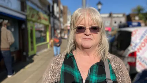 Sam Dixon-French / BBC A woman wearing sunglasses stood on a pavement near parked cars. She is weating a knitted cardigan and a green and black tartan top.