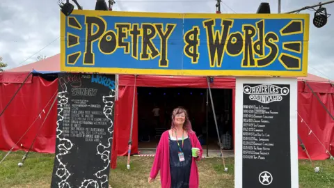 Sally Jenkinson wearing pink top and black outfit, stood in front of huge blue and yellow sign saying 'Poetry & Words'.