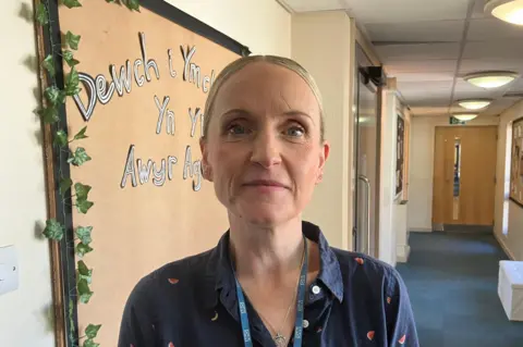 A woman with pulled back blonde hair wearing a blue shirt with a watermelon slice pattern, stands in a school corridor. She is wearing a lanyard with an NHS logo ribbon.