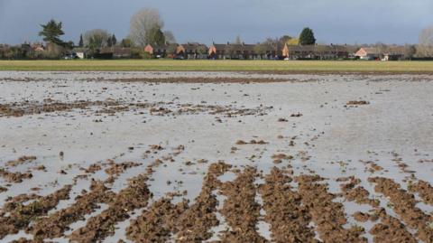 A ploughed field containing channels of water, with houses in the background