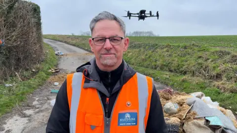 Carl McIvor, waste crime manager at Maidstone Borough Council, pictured near Yalding, Kent, with the drone flying in the background. Fly-tipped items can be seen in the background, and Carl is wearing a black fleece and orange high-vis jacket. He has graying hair and glasses.