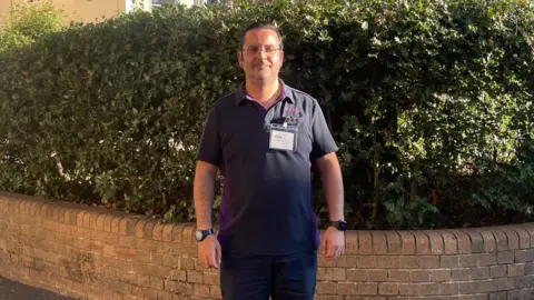 A man in glasses stands in front of a low wall in the street. He is wearing a dark polo shirt and dark-coloured trousers. He is smiling.
