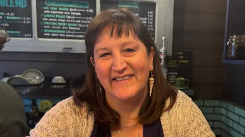 BBC Helen smiling behind counter. She has a fringe and long, dark hair reaching her shoulders and is wearing a cream crochet cardigan. One earring is made from a long spiral seashell while the other is long, silver and wavy. A menu can be seen on the wall behind her. 