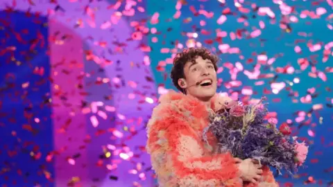 Reuters An elated-looking Nemo - the Swiss singer who won Eurovision in 2024. He has short, dark, curly hair, is wearing a coral pink fluffy coat and holding a large bunch of flowers. He is surrounded by falling sparkling confetti.