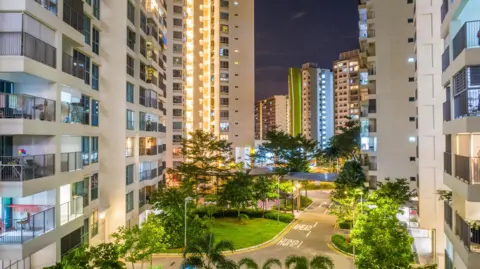 Getty Images Singapore city centre at nigh, there are several high-rise flats either side of a road and they are light in colour with railed balconies. In between the buildings is a road with "ahead hump" written on it, as well as yellow cross hatches. There are several trees and a small patch of grass separating the buildings as well 