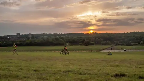 Getty Hainault Forest Country Park at sunset with the sun glowing orange behind a line of distant trees and low hills. In the foreground, three children move across the open space: one running on foot, one riding a bicycle, and another sitting beside a bicycle. The sky is filled with soft, layered clouds reflecting warm evening light, while a few distant buildings and a parking area sit at the far edge of the landscape.