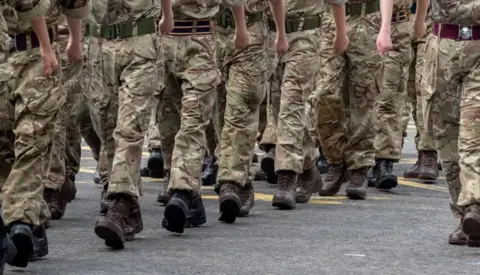 Legs, the identity of each person obscured, walk in a row with camouflaged trousers and black Army boots