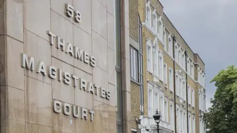 Two police officer in a high-visibility yellow jackets stand in the foreground, at the the entrance of Thames Magistrates' Court. The building's stone façade features the court's name in large, metallic lettering below the number 58. To the right, a traditional brick building with white window frames and a black lamp post lines the street.