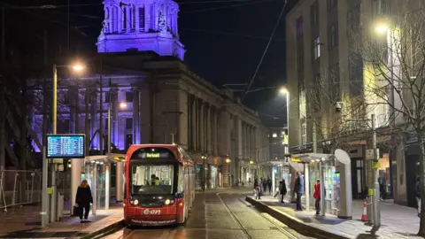 Night time view of the Old Market Square in Nottingham. In the foreground, a red tram stopped at the station, with several passengers waiting on the platforms. In the background, the Council House, lit up by blue lights.