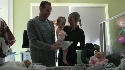 Liam (left) stands in a hospital room holding a stuffed unicorn, showing it to Sienna. Natasha stands next to him holding Sienna.