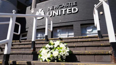 A white and green floral tribute on concrete steps outside of St James' Park. At the top of the stairs there are glass doors against a dark grey black building with 'Newcastle United' written in white lettering above. The club crest is to the left.
