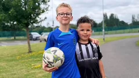 Two children wearing football jerseys pose proudly for the camera. The child on the left holds a football.