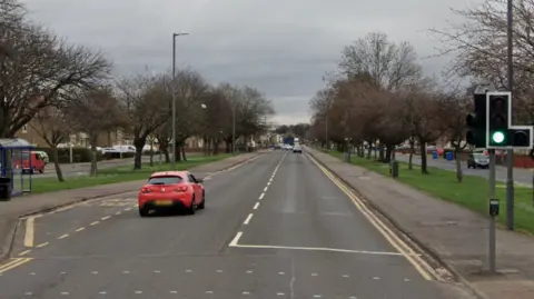 An urban road lined with trees. It has a pedestrian crossing and a bus stop.