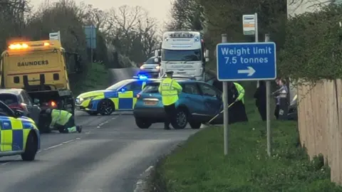 Paul Pinkstone A police car and ambulance attend a road incident where a large box trailer has overturned, blocking both lanes of a rural road.