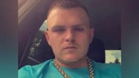 A man with short hair and wearing a gold chain and a green T-shirt is sitting in a car.