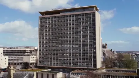 The picture shows a tall, rectangular tower with a grid of small square windows running across its entire front. The building looks weathered and empty, with many of the windows appearing dark or discoloured. It stands above a cluster of lower, flat‑roofed concrete buildings that look like part of the same complex. To the right, behind the tower, there is an older stone building with pointed rooftops and spires peeking through the trees. In the distance, you can see more low‑rise buildings, patches of greenery and the outline of rolling hills under a bright sky filled with scattered clouds.