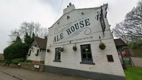 Front exterior of The British Oak pub, a white-painted brick building with large “Ale House” lettering on the façade, hanging baskets and a roadside setting bordered by trees.