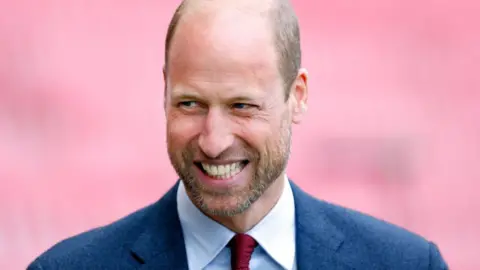 Prince William, wearing a navy suit and red tie, smiles
